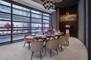 Un área de comedor moderna con una mesa grande dispuesta para seis personas, rodeada de sillas, y vista a un centro comercial a través de amplios ventanales.