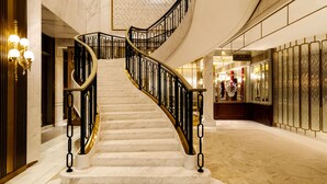Una gran escalera de mármol con barandilla negra que conduce a un vestíbulo espacioso con techos altos y un candelabro decorativo.