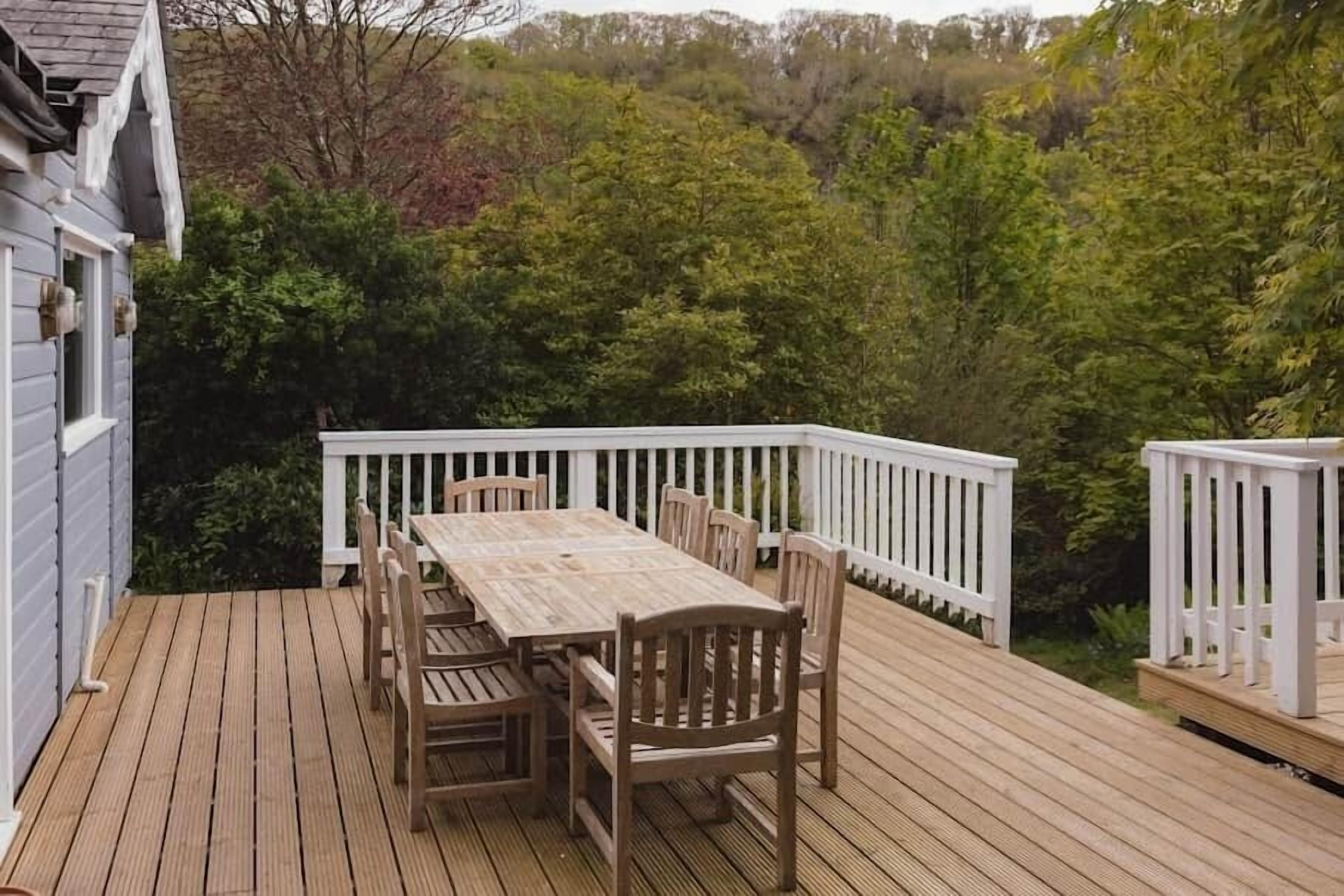 A wooden deck with a table and chairs, surrounded by white railings and dense greenery.