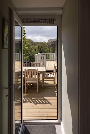 A view through a glass door to an outdoor deck with a table and chairs, surrounded by greenery.