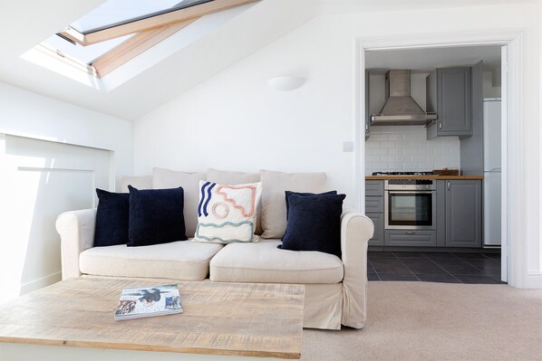 A modern living room with a sofa, pillows, and a coffee table. There is a skylight above the sofa and a kitchen visible through an open doorway. - Spacious 3 bedrooms flat with terrace (London)