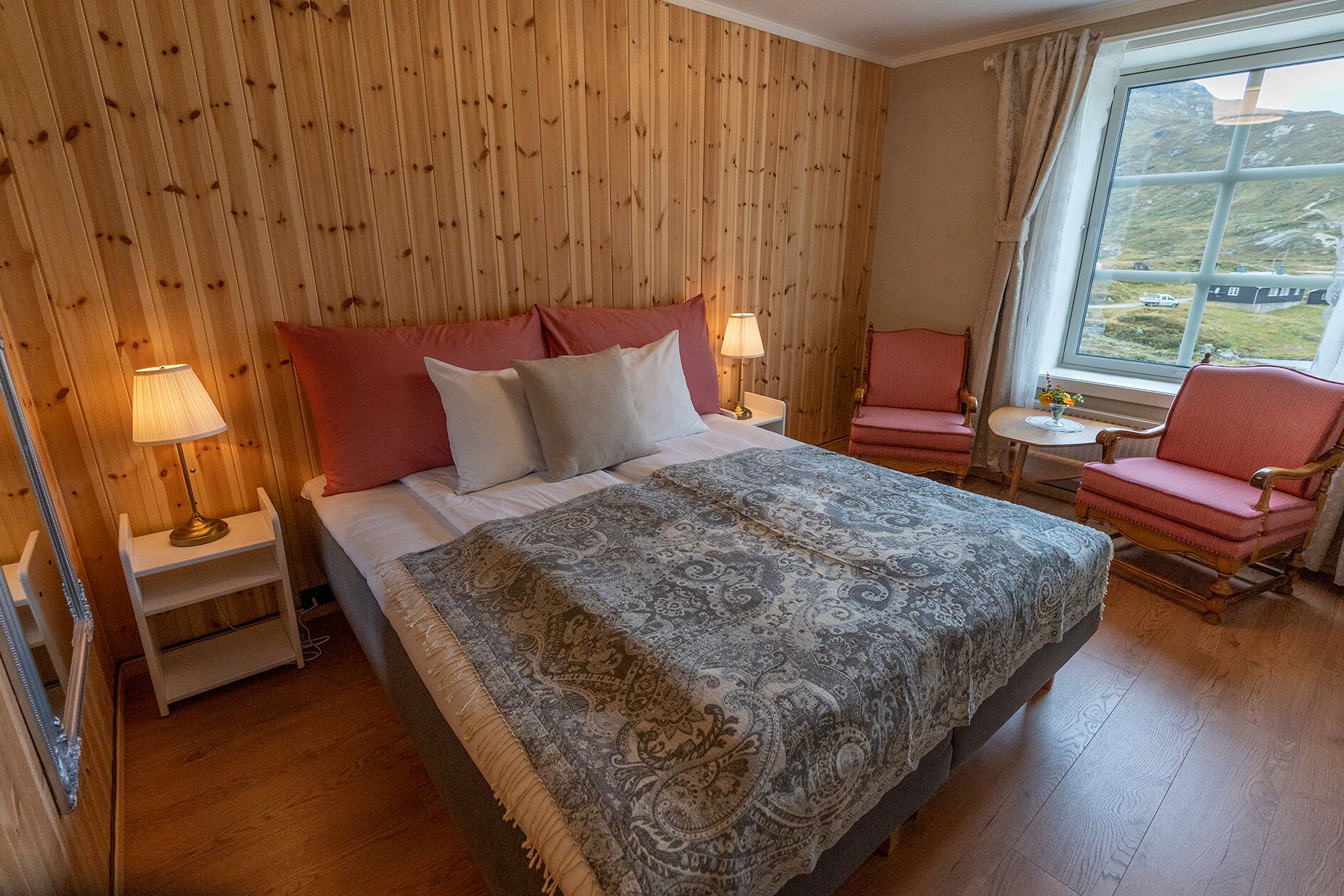 Un dormitorio acogedor con un panel de madera, una cama grande con ropa de cama estampada, dos lámparas de noche, una mesita con un florero y una silla junto a la ventana.