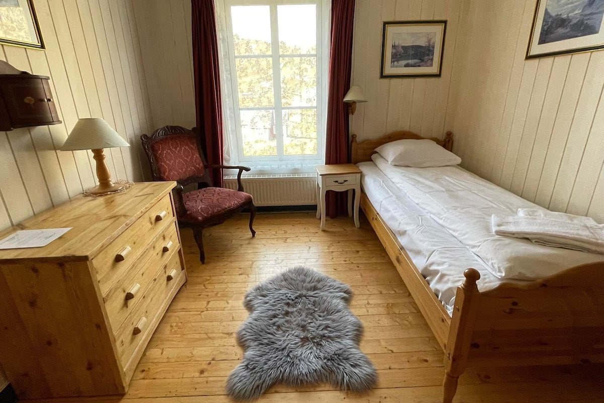 Une chambre avec une commode en bois, une chaise, un lit, une table de chevet, une lampe, une fenêtre avec des rideaux et un tableau encadré au mur.