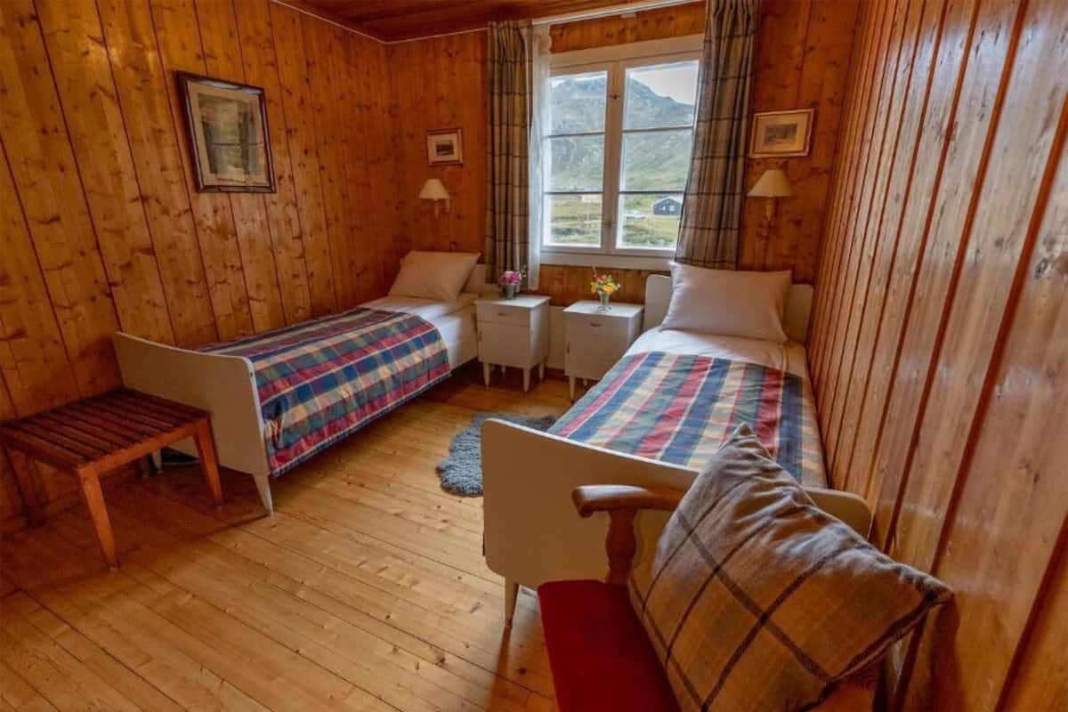 Une chambre rustique en bois, avec deux lits, un banc en bois, une petite table et une fenêtre donnant sur les montagnes.