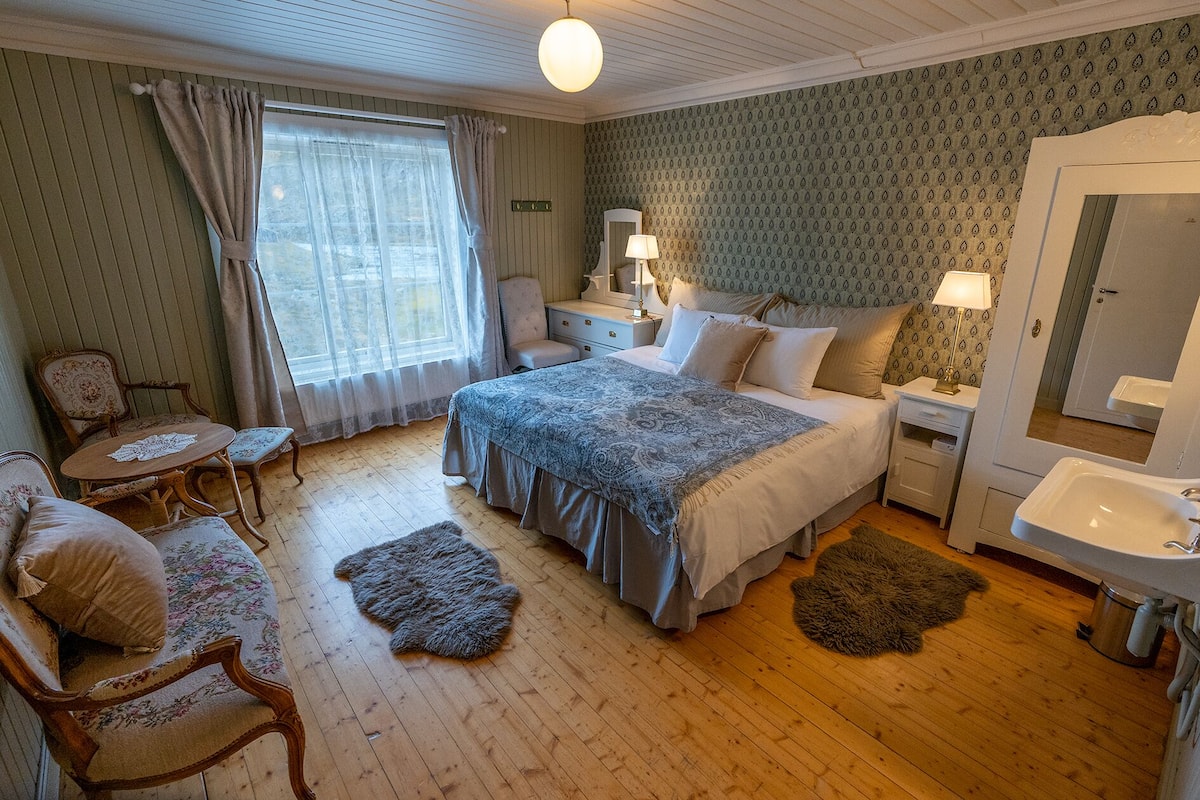 Une chambre confortable avec un grand lit, un coin salon avec un fauteuil et une table, une commode, un miroir et une salle de bain avec un lavabo.