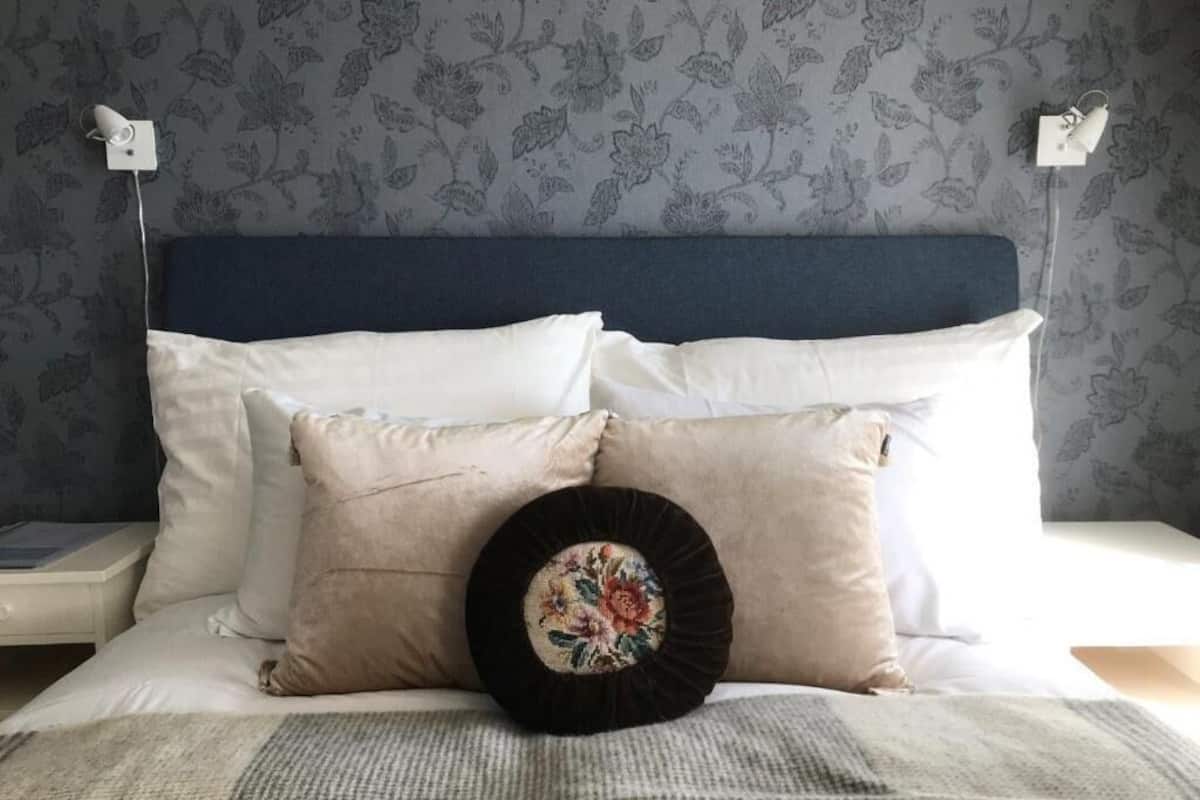Un lit bien fait, avec des oreillers blancs et beiges, un coussin noir décoratif orné d’un motif floral, et une tête de lit bleue sur un fond de papier peint à motifs floraux.