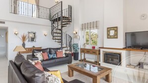A spacious living room with a black leather sofa, a wooden coffee table, and a fireplace. A spiral staircase leads to an upper level. - La Manga Club Los Olivos Apartment 593 (La Manga Club, Cartagena)