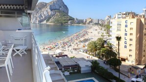 Ein Balkon mit weißen Stühlen, einem Pool und Blick auf einen Strand mit Sonnenschirmen und Menschen.