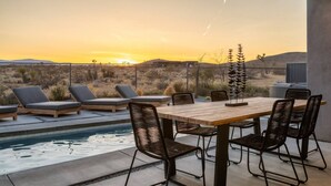 A poolside area with a wooden table and chairs, lounge chairs, and a decorative plant.
