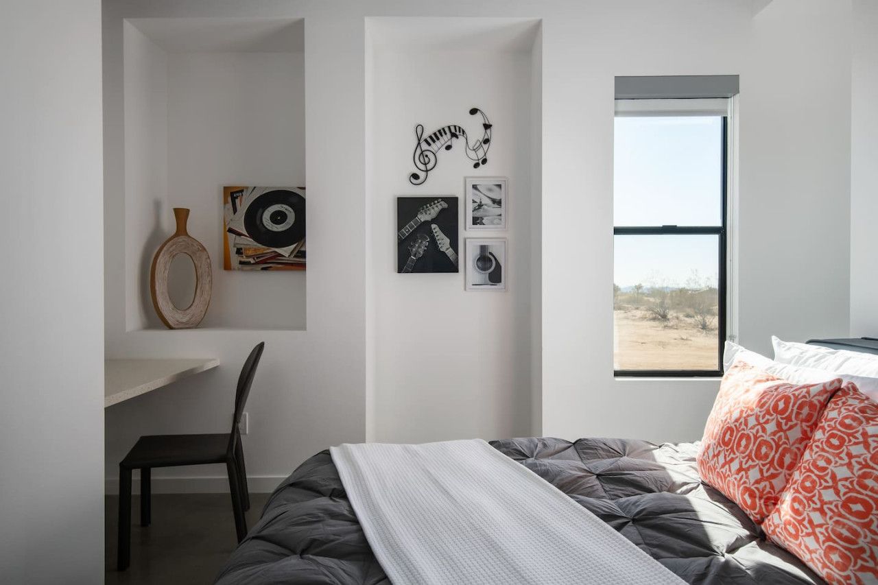 Ein Schlafzimmer mit Bett, Schreibtisch und Stuhl. An den Wänden befinden sich Kunstwerke und ein Fenster mit Blick.