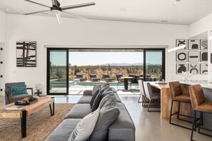 A modern living room with a large gray sofa, a coffee table, and a dining area with bar stools. A large sliding glass door leads to an outdoor patio with a pool and chairs.