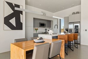 A modern kitchen with a central island, a dining area with a wooden table and chairs, and a large abstract painting on the wall. - W House: Luxe • Near Park • Heated Pool & Spa (Joshua Tree)