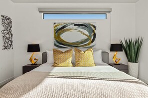 A modern bedroom with a large bed, two bedside tables with lamps, a large abstract painting above the bed, and a potted plant on the right.