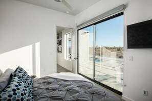 A bedroom with a large sliding glass door leading to a pool area, a bed with a grey quilt, and a wall-mounted TV. - W House: Luxe • Near Park • Heated Pool & Spa (Joshua Tree)
