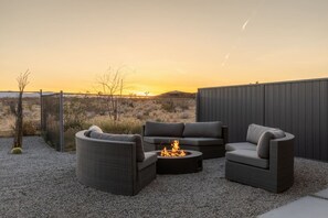 An outdoor seating area with a fire pit, surrounded by a gravel patio and a view of the desert landscape at sunset. - W House: Luxe • Near Park • Heated Pool & Spa (Joshua Tree)