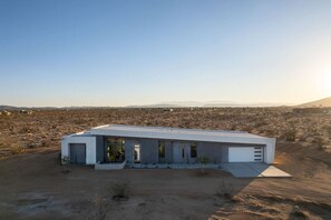 A modern, single-story building with a flat roof, large windows, and a minimalist design, situated in a desert landscape with distant mountains. - W House: Luxe • Near Park • Heated Pool & Spa (Joshua Tree)