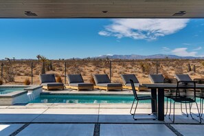 A pool area with lounge chairs and a table, surrounded by a desert landscape with mountains in the background. - W House: Luxe • Near Park • Heated Pool & Spa (Joshua Tree)