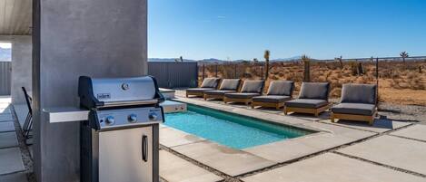 Un espace aménagé au bord de la piscine, comprenant un barbecue, des chaises longues et une vue sur le paysage désertique.