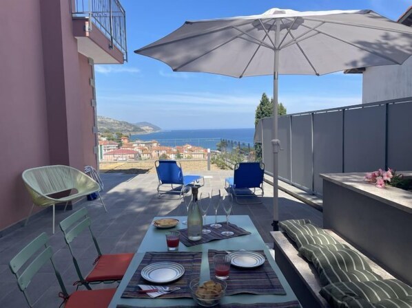 A terrace with a table set for two, a large umbrella, and a view of the sea.
