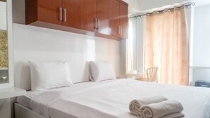 Una cama bien tendida con sábanas blancas y dos toallas enrolladas, un armario de madera arriba y una ventana con cortina.
