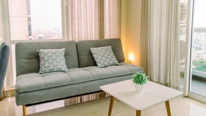 A grey sofa with patterned cushions, a white coffee table with a plant, and a city view through sheer curtains. - Elegant And Restful 2Br At Menteng Park Apartment (Jakarta)