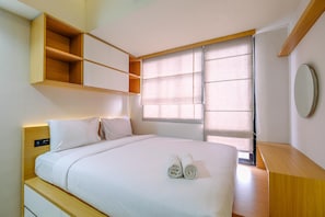 A neatly made bed with two rolled towels, a wooden bedside table, a built-in shelf unit, and a window with blinds. - Best Deal And Tidy Studio At Evenciio Margonda Apartment (Depok)