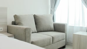 A modern, minimalist living room with a grey sofa, a small side table, and a large window with sheer curtains.