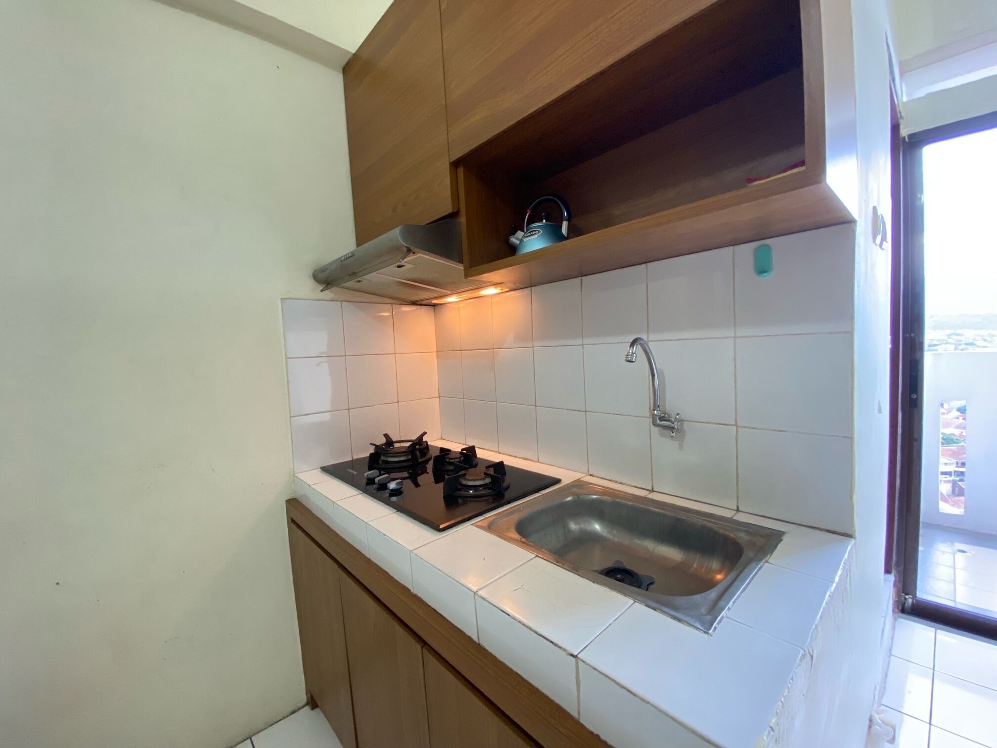 A kitchen with white tiles, a stainless steel sink, a gas stove, wooden cabinets, and a window with a view.
