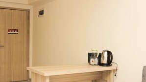 Ein Holztisch mit einem Wasserkocher und einer Flasche, ein grauer Stuhl und eine Holztür mit einem Schild.