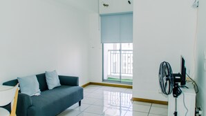 A room with a grey sofa, a small table, a standing fan, and a window with a view of a balcony. - Fancy And Nice 2Br Apartment At M-Town Residence (Gading Serpong)