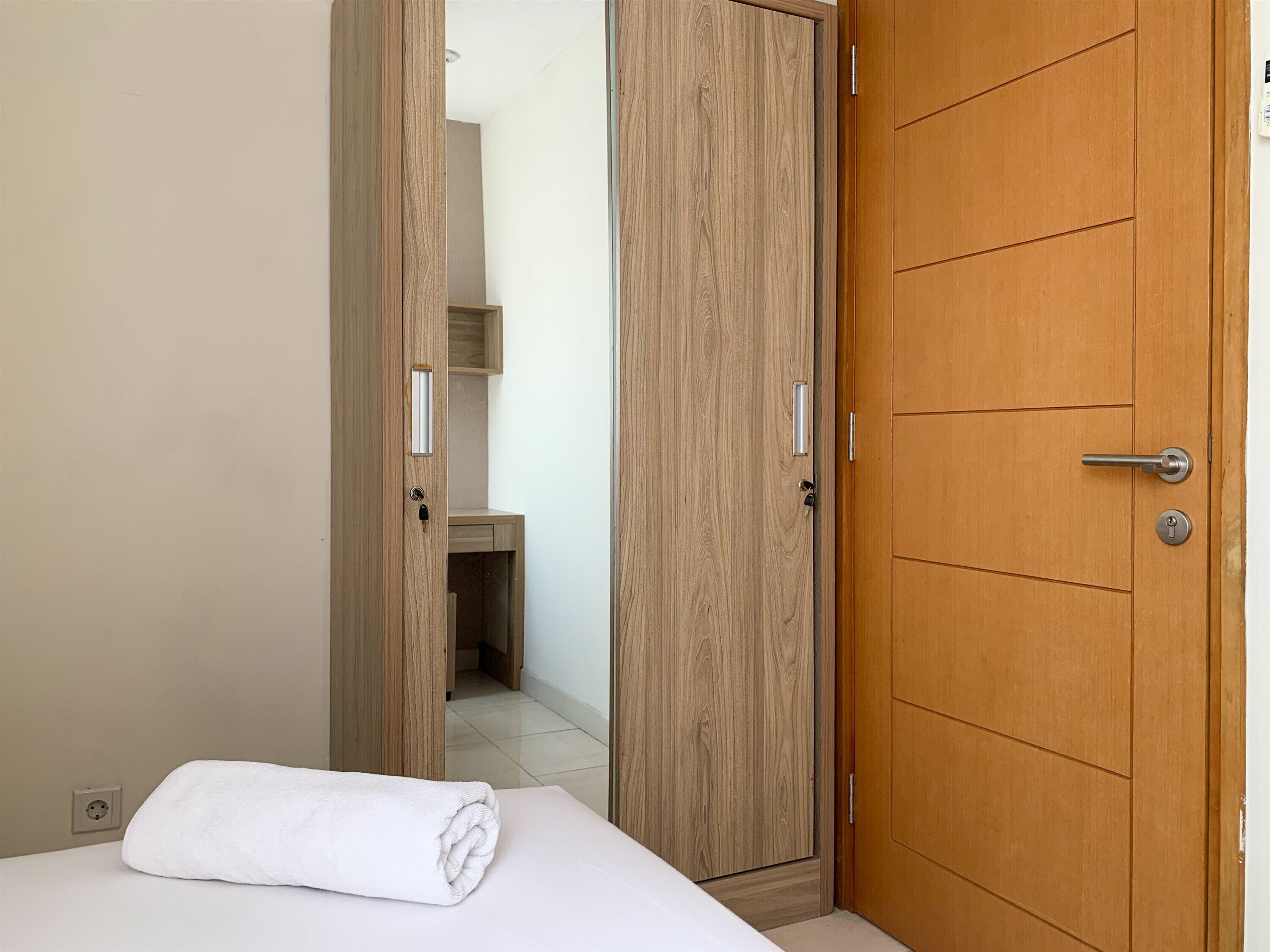 A hotel room with a wooden wardrobe, a bed with a white towel, and a small wooden shelf.