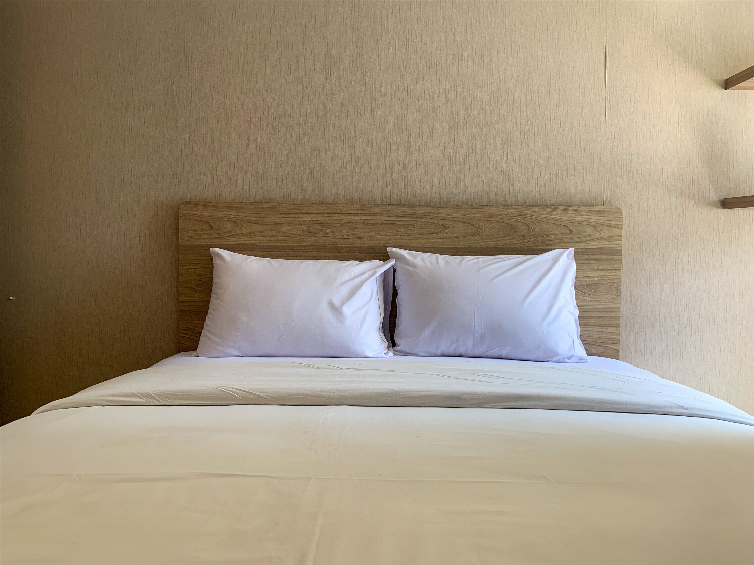 Un lit bien fait, avec du linge de lit blanc et deux oreillers, adossé à une tête de lit en bois et placé devant un mur de couleur beige.