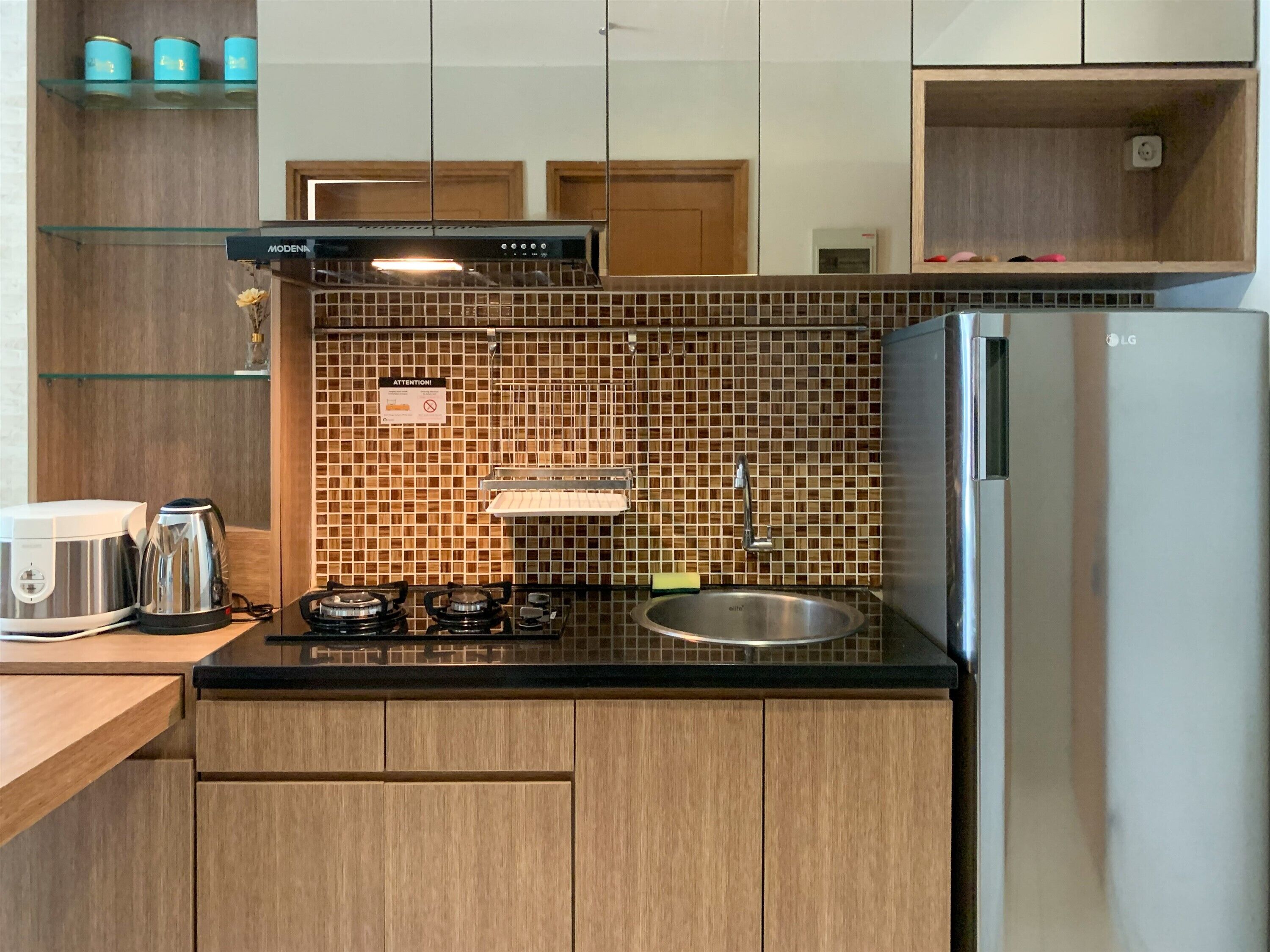 A compact kitchen with a gas stove, a stainless steel sink, and a refrigerator. There are wooden cabinets and a tiled backsplash.
