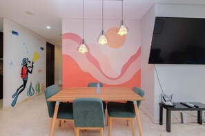 A dining area with a wooden table, green upholstered chairs, pendant lights, a wall mural, and a flat-screen TV.