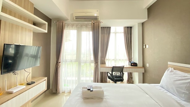 A hotel room with a bed, a desk, a chair, a TV, and an air conditioning unit. - Cozy Studio Room At Taman Melati Jatinangor Apartment (Hegarmanah)