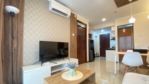 A modern living room with a flat-screen TV, a coffee table with a vase, a dining area with a white table and chairs, and a kitchen area with a refrigerator and microwave.