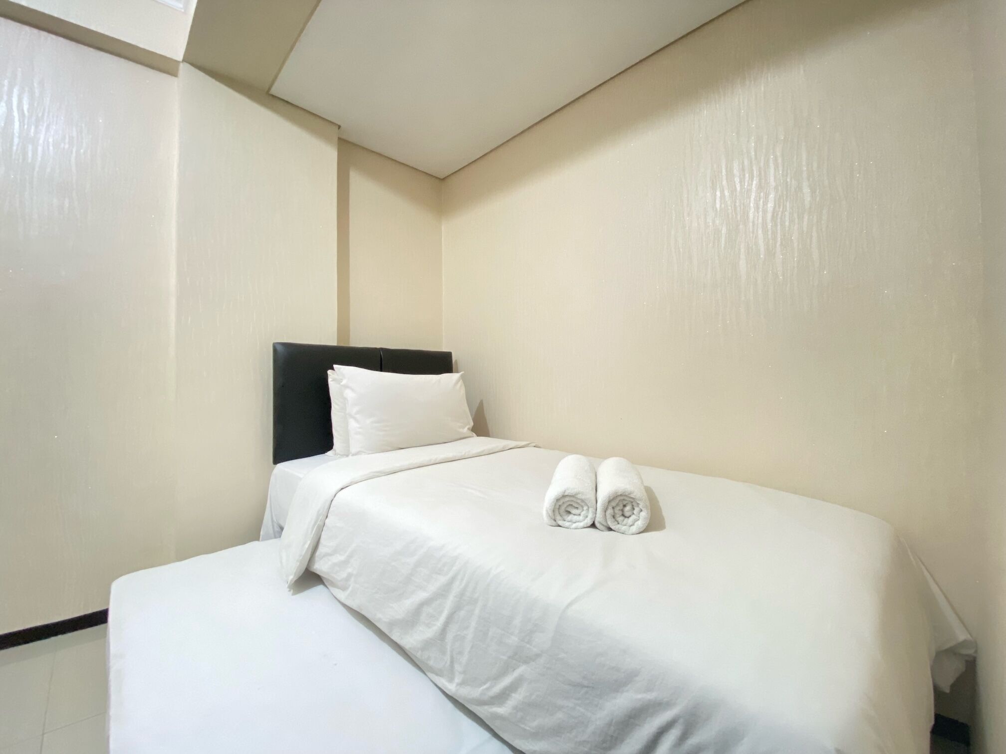 A hotel room with a bed, white bedding, a black headboard, and a pair of rolled towels on the bed.