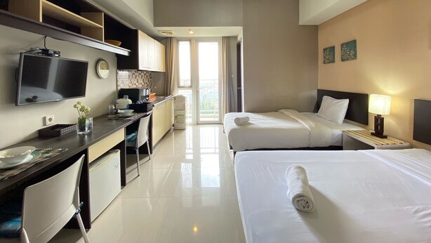 A hotel room with a flat-screen TV, a desk with a computer, two beds, a nightstand with a lamp, and a view of the outdoors through a door. - Cozy Studio Room At Skyland City Jatinangor Apartment (Hegarmanah)