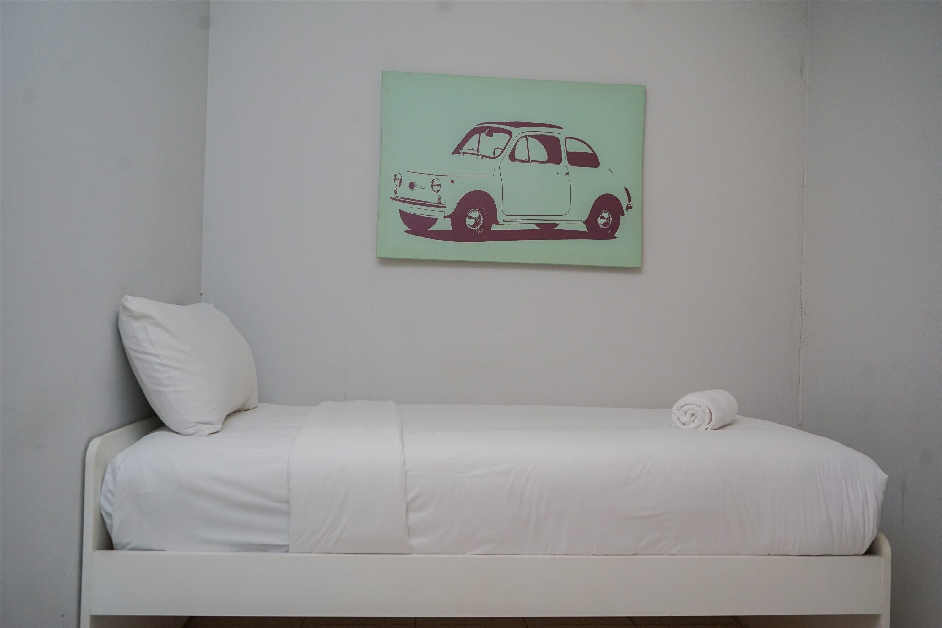 Una cama individual con ropa de cama blanca y un almohadón, una pared verde con ilustración de un coche, y una puerta blanca.