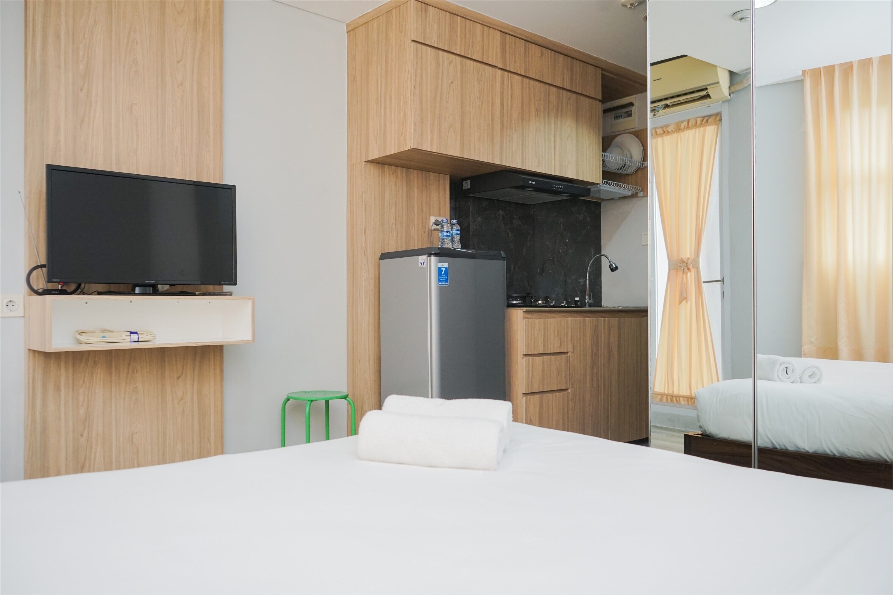 A hotel room with a bed, a TV, a kitchenette with a fridge, and a green stool.