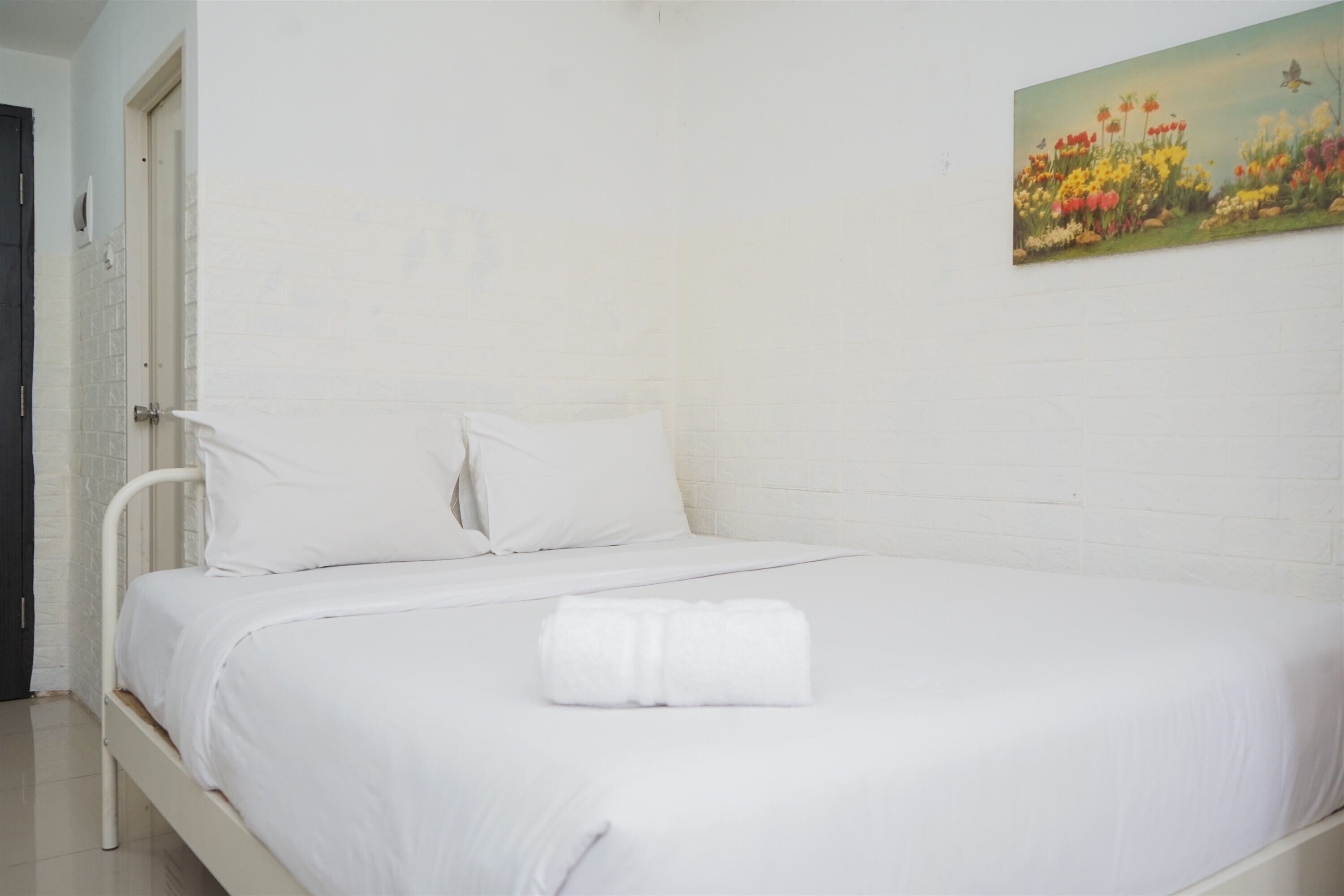 Un lit bien fait, avec du linge de lit blanc et une serviette enroulée, un mur en briques blanches et un tableau représentant des fleurs accroché au mur.