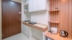 A compact kitchen with a wooden door, white cabinets, a countertop with a kettle and a rice cooker, and a small shelf with decorative items.