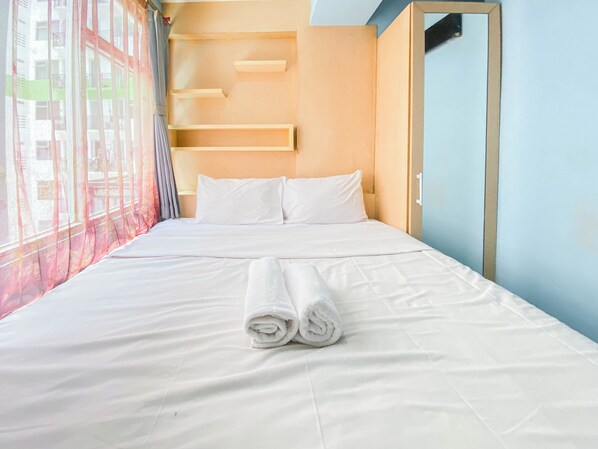 Un lit bien fait, avec du linge de lit blanc et des serviettes pliées, une fenêtre ornée de rideaux légers et un mur bleu clair agrémenté d’une étagère en bois.