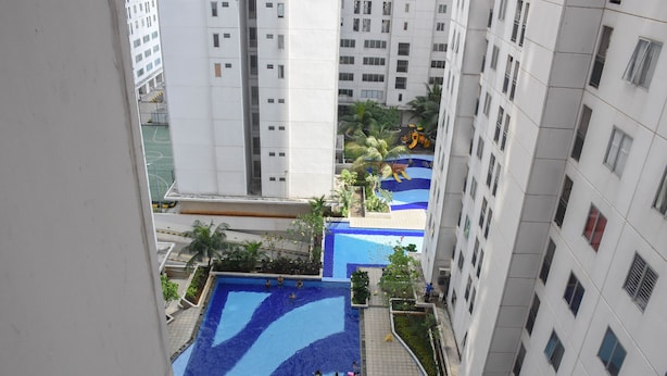 A high-rise building with a swimming pool area featuring blue wave patterns, surrounded by greenery and palm trees. - Enjoy Living And Tidy 1Br Bassura City Apartment (Jakarta)