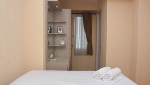 A hotel room with a neatly made bed, a small shelf with a clock and picture, and a window with curtains. - Enjoy Living And Tidy 1Br Bassura City Apartment (Jakarta)