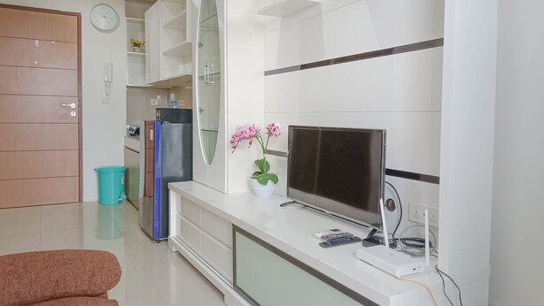 A modern living room with a flat-screen TV, a white entertainment unit, a glass cabinet, and a potted plant. - Nice And Homey 2Br At Vida View Makasar Apartment (Makassar)