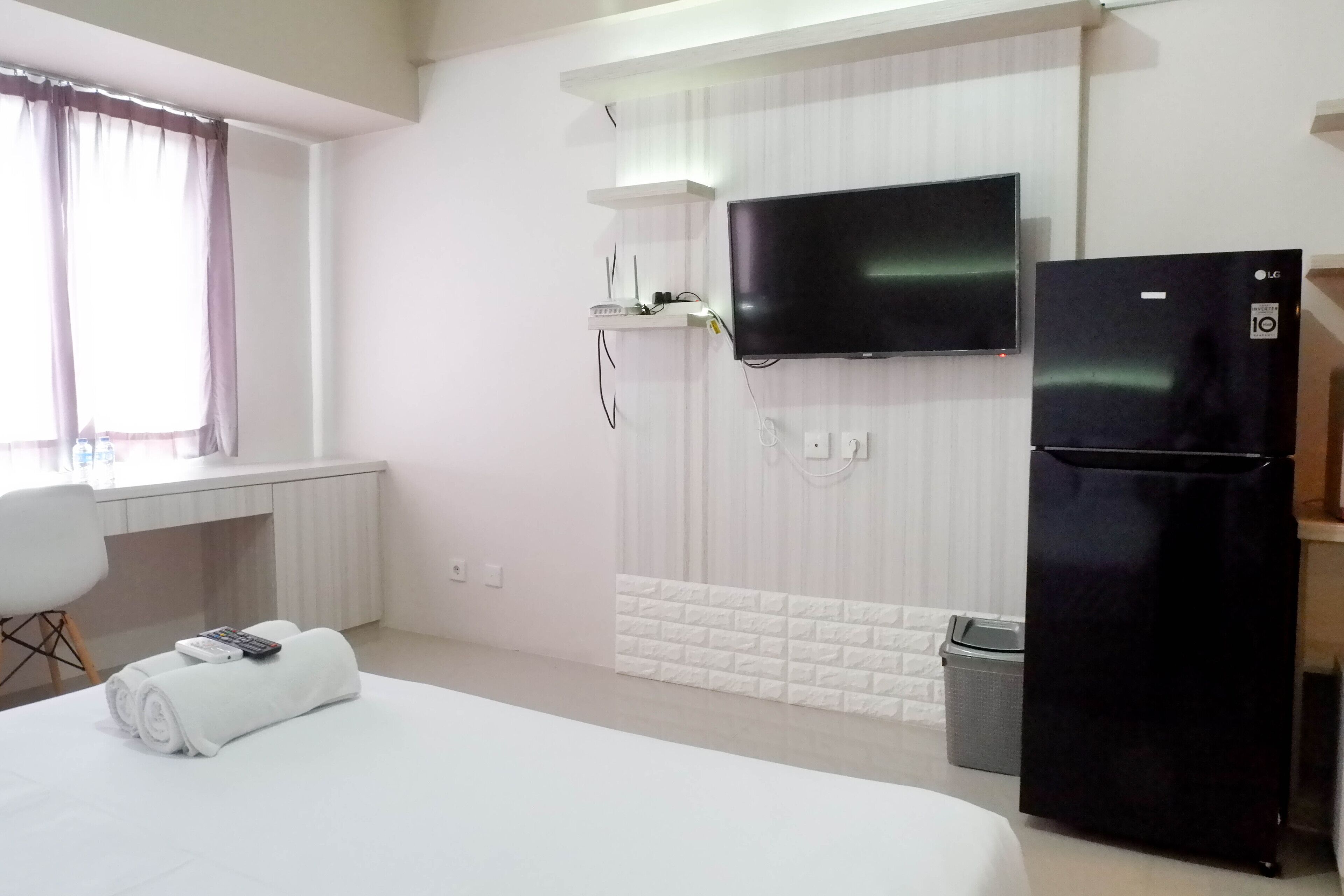 A hotel room with a bed, a TV mounted on the wall, a refrigerator, a desk with a chair, and a window with curtains.