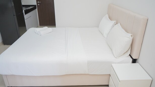 A neatly made bed with white bedding and pillows, a small bedside table, and a kitchen area visible in the background. - Nice And Modern Studio At Transpark Bintaro Apartment (South Tangerang)