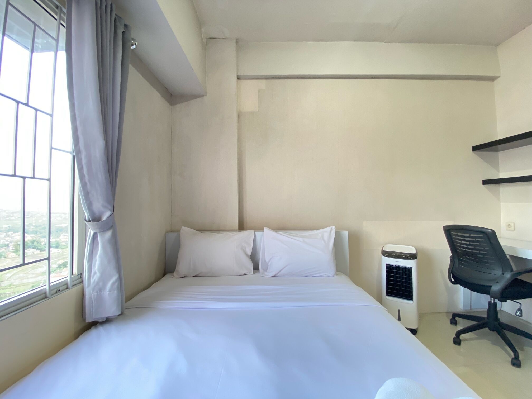 Habitación de hotel con una cama, un escritorio y una silla. Se ve una ventana con cortinas, una pared blanca y un respiradero en el techo.