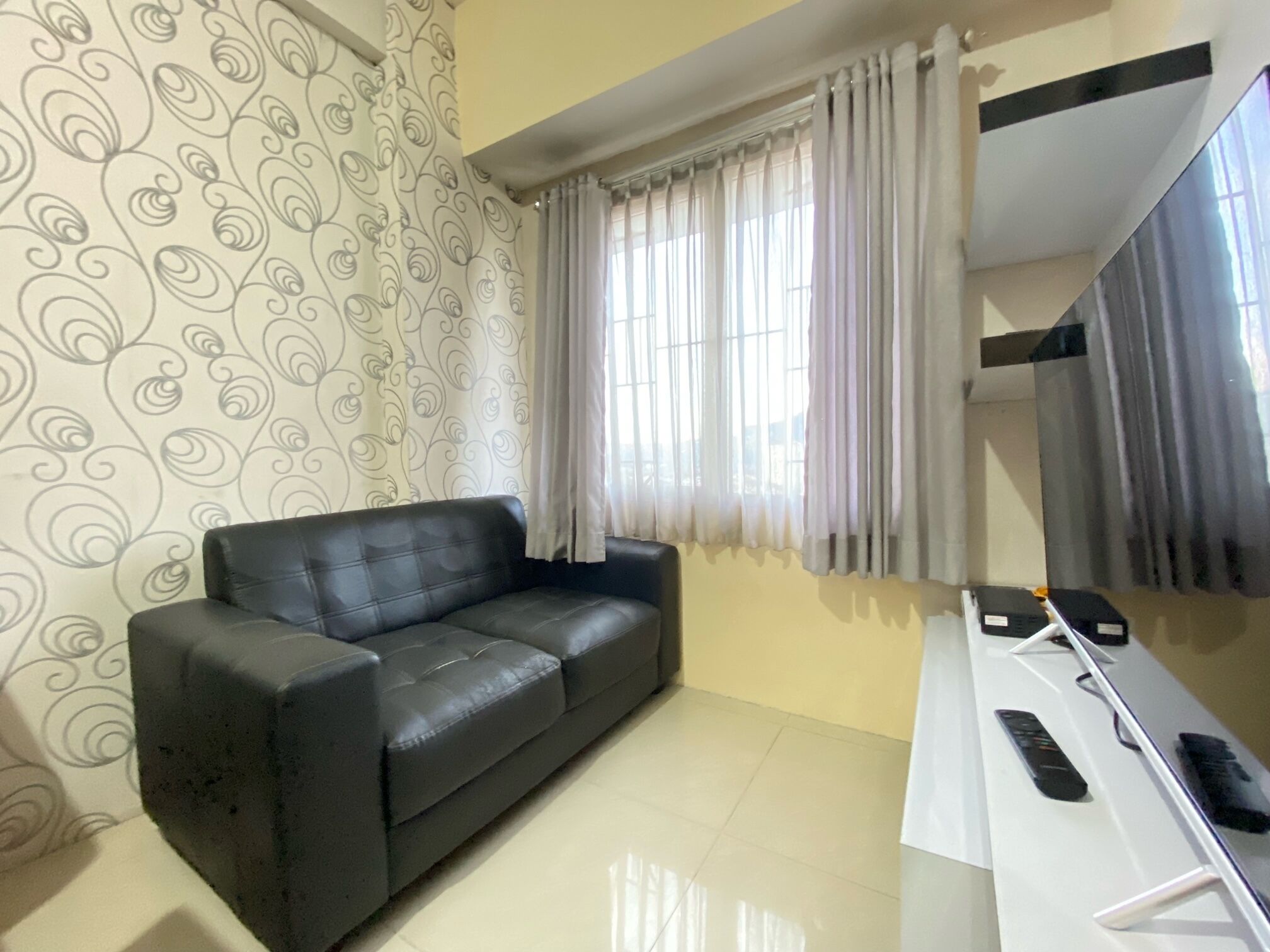 Una habitación con un sofá de cuero negro, un escritorio blanco con controles remotos y una ventana con cortinas traslúcidas.